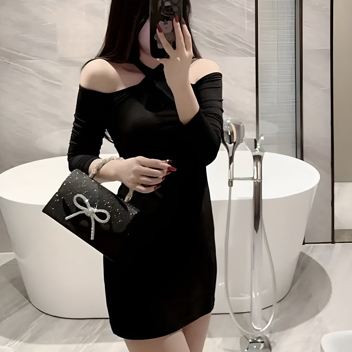 Person wearing a black off-shoulder dress holding a black handbag with a bow in a bathroom setting.